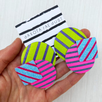 A pair of oversize stripey studs made of a selection of geometric shapes mounted on a dakota rae dust branded card, held in a woman's hand
