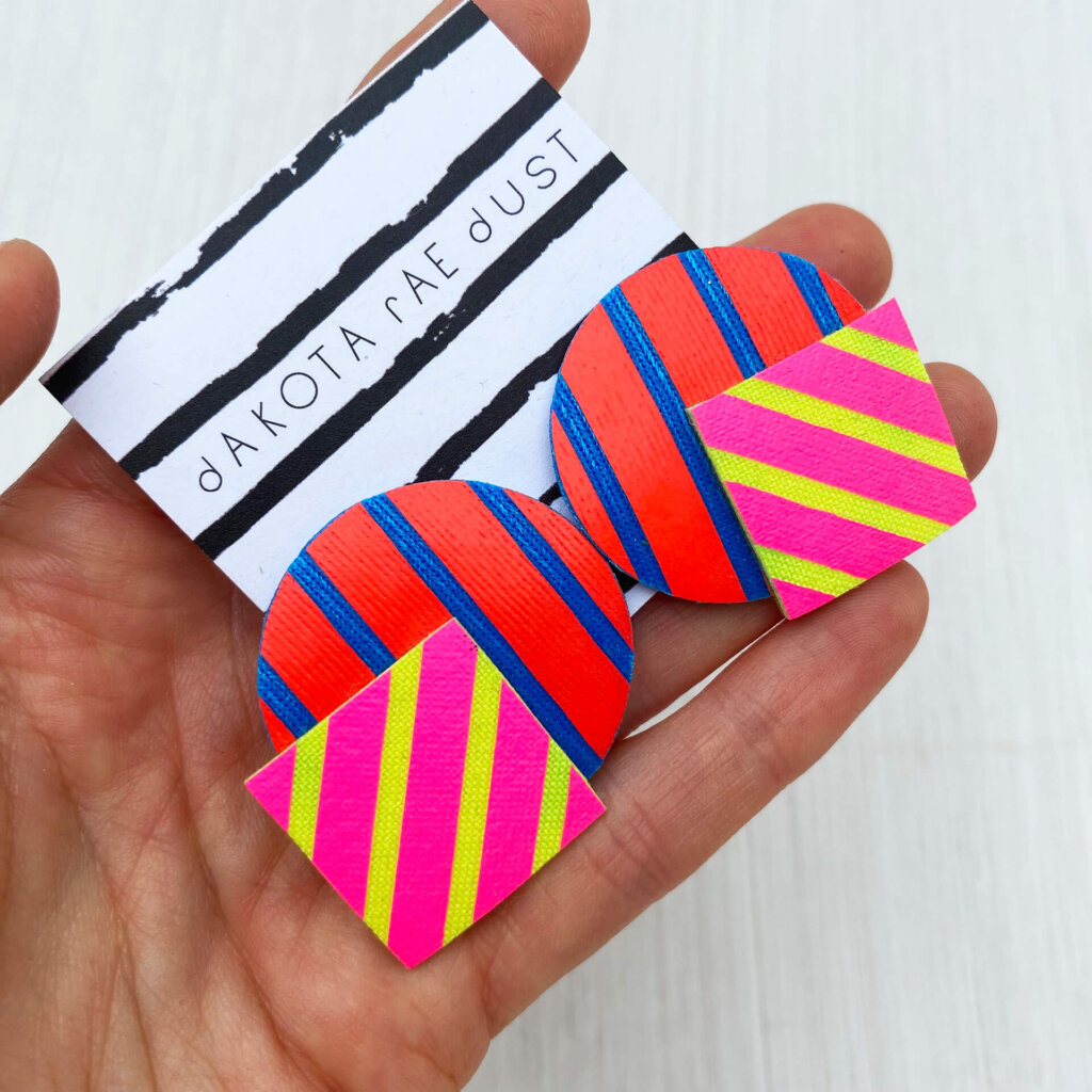 A pair of oversize stripey studs made of a selection of geometric shapes mounted on a dakota rae dust branded card, held in a woman's hand