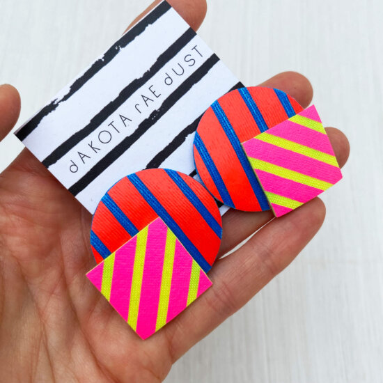 A pair of oversize stripey studs made of a selection of geometric shapes mounted on a dakota rae dust branded card, held in a woman's hand