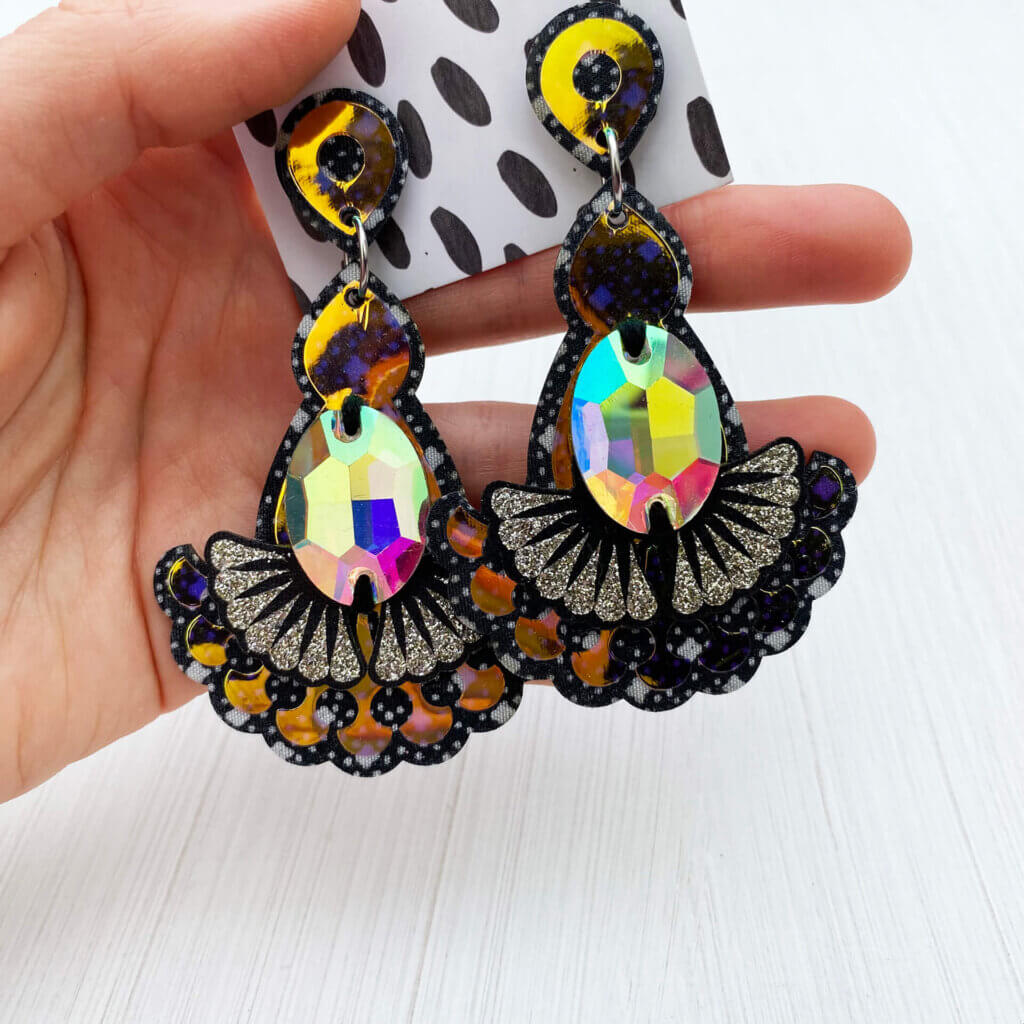 A pair of sparkly iridescent jewel earrings is held in an open hand in front of a plain white background