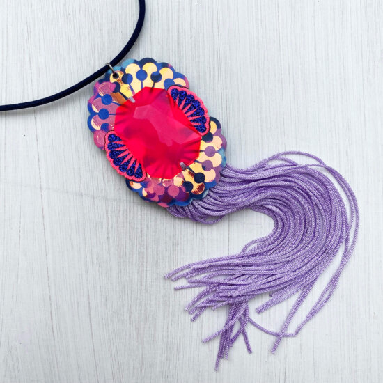 A neon pink, navy and lilac custom cast giant jewel pendant necklace with a lilac fringe lying on an off white background