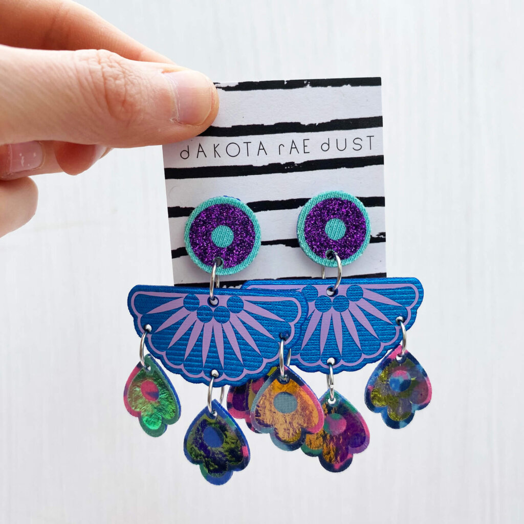 A pair of colourful chandelier style, jangly droplet earrings mounted on a black and white striped, dakota rae dust branded card are held between a just visible thumb and forefinger