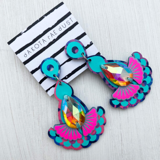 A pair of turquoise and pink jewel earrings mounted on a black and white striped, dakota rae dust branded card are lying on a plain off white background