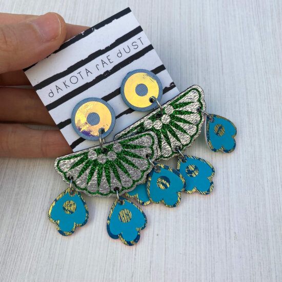 A pair of colourful chandelier style, jangly droplet earrings mounted on a black and white striped, dakota rae dust branded card are held between a just visible thumb and forefinger