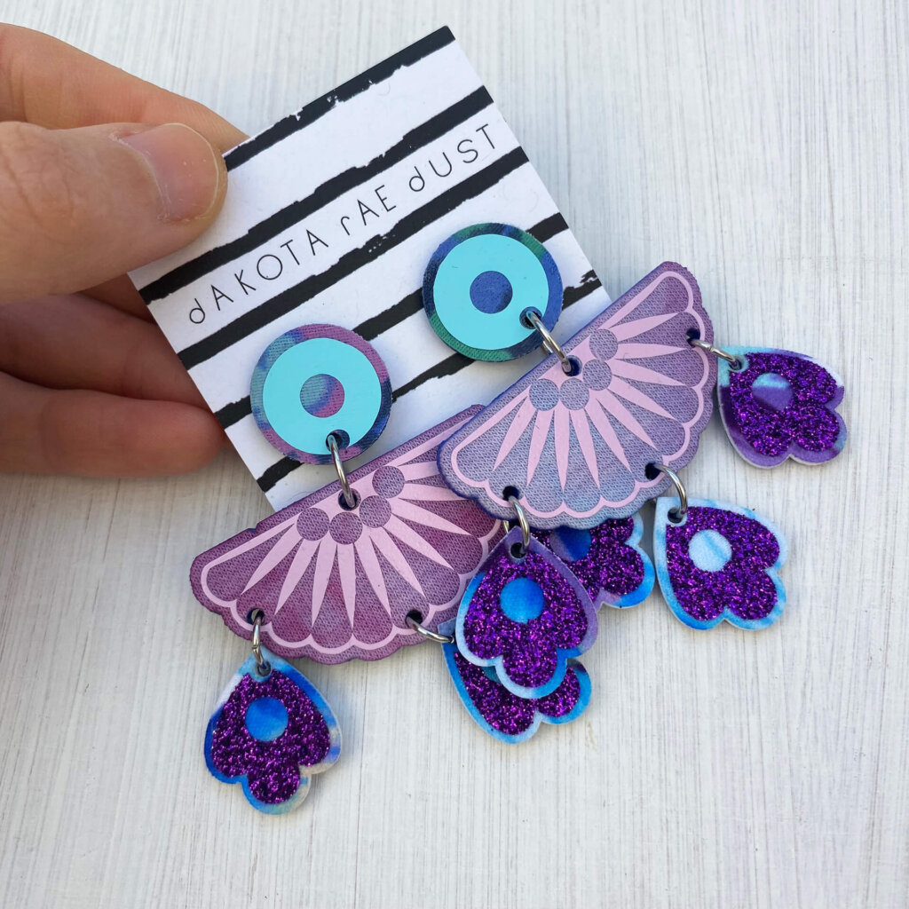 A pair of colourful chandelier style, jangly droplet earrings mounted on a black and white striped, dakota rae dust branded card are held between a just visible thumb and forefinger