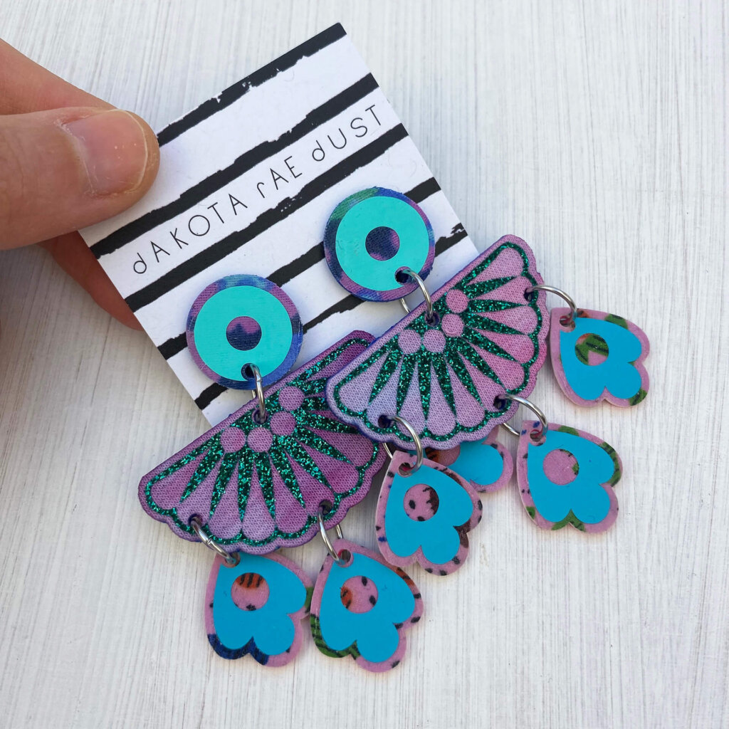 A pair of colourful chandelier style, jangly droplet earrings mounted on a black and white striped, dakota rae dust branded card are held between a just visible thumb and forefinger