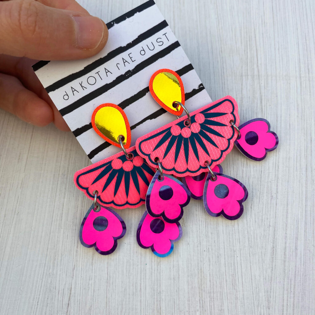 A pair of colourful chandelier style, jangly droplet earrings mounted on a black and white striped, dakota rae dust branded card are held between a just visible thumb and forefinger