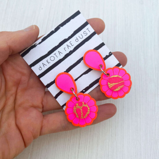 A close up of pair of neon pink small flower earrings mounted on a black and white patterned dakota rae dust branded card held in an open hand