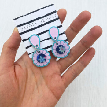 A close up of pair of lilac small flower earrings mounted on a black and white patterned dakota rae dust branded card held in an open hand