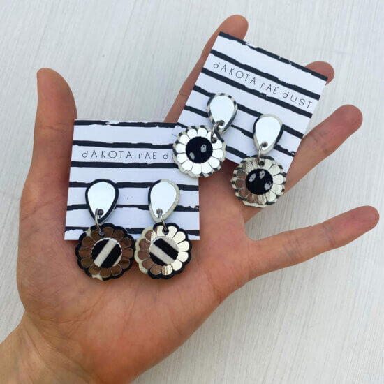 Two pairs of small flower earrings mounted on a black and white patterned dakota rae dust branded cards are held in an open hand