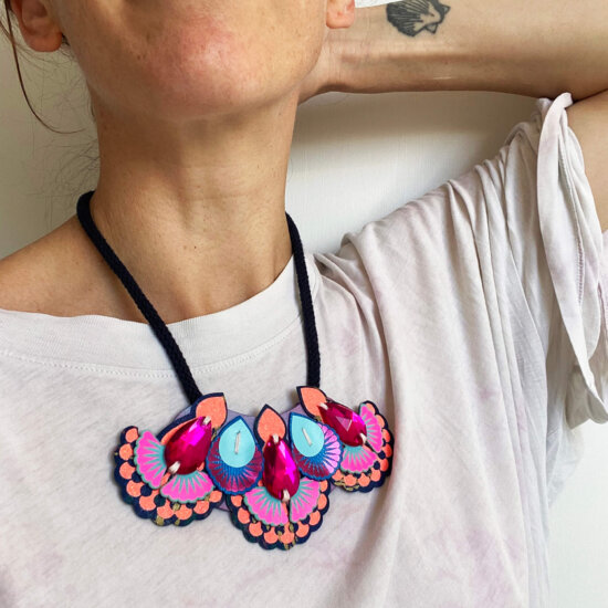 A close up of th eneck and chest area of a woman wearing a loose fit off white t-shirt and colourful mini bib necklace. Her left arm is raised and her hand is behind her head.