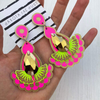 A pair of gold and neon jewel earrings are held in an open hand