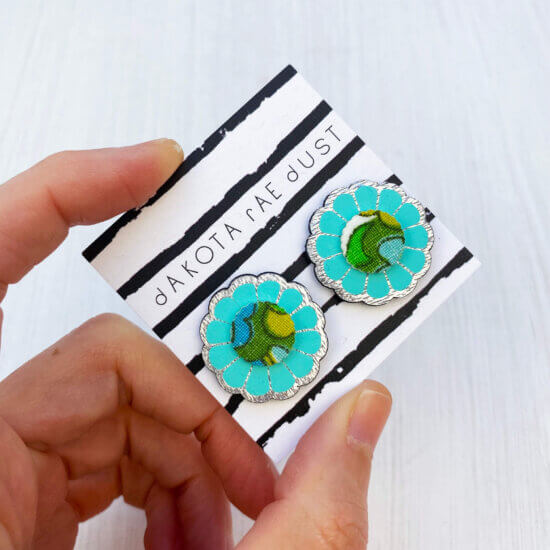 A pair of turquoise green summery flower studs mounted on a dakota rae dust branded card are held against an off white background in a just visible thumb and forefinger.